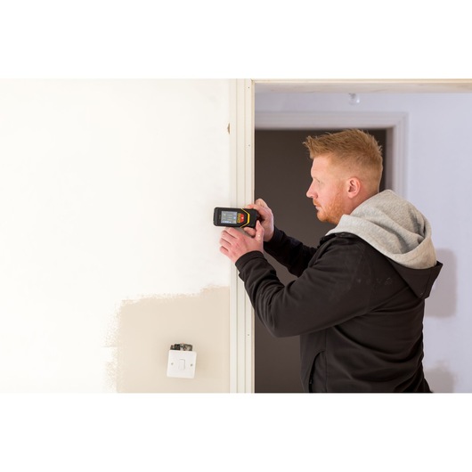 Foto van STANLEY STHT1-77139 elektronische detector gebruikt op muur, persoon in zwarte hoodie bedient apparaat.