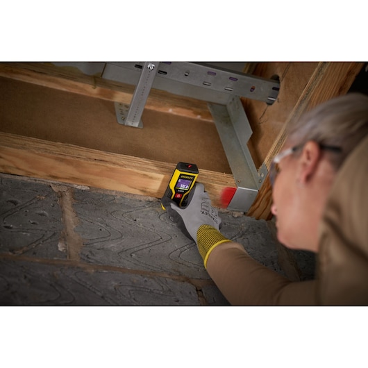 STANLEY STHT1-77409 télémètre laser tenu par une main gantée mesurant une poutre en bois à l'intérieur.