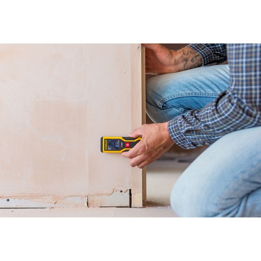 Télémètre laser STANLEY STHT1-77409 utilisé contre un mur pendant une mesure, utilisateur en tenue de travail décontractée.