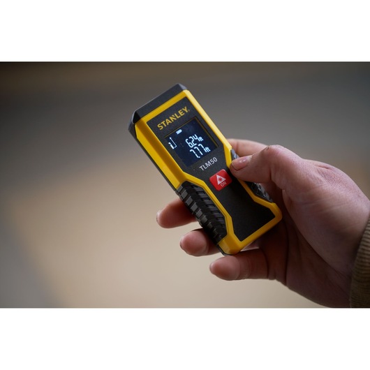 Photo du télémètre laser STANLEY STHT1-77409 tenu à la main, jaune et noir avec écran numérique.