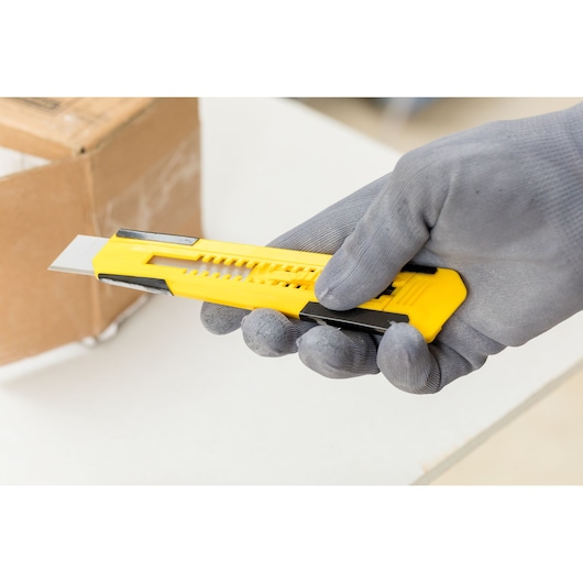STANLEY STHT10345-0 hobbymes met gele handgreep, vastgehouden door een gehandschoende hand, doos op de achtergrond.