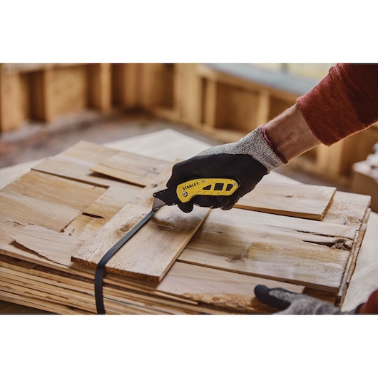 Couteau pliant STANLEY STHT10424-0 coupant une sangle noire sur du bois empilé, vue rapprochée.