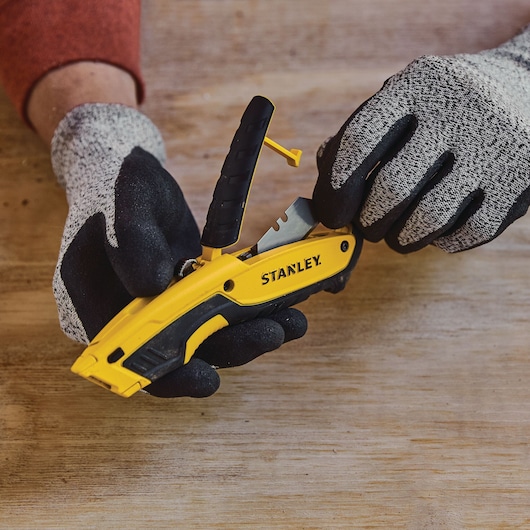 Photo du couteau utilitaire jaune STANLEY STHT10479-0 avec poignée noire tenu par des mains gantées sur une surface en bois.