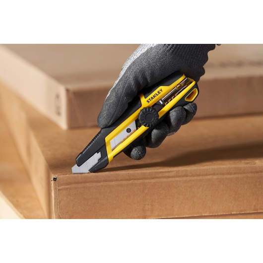 Foto van STANLEY STHT10500-0 afbreekmes met geel-zwarte handgreep, karton snijdend, vastgehouden door gehandschoende hand.