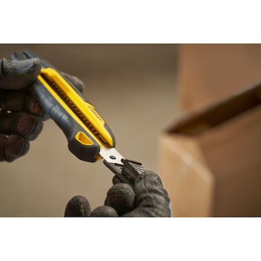 Stanley STHT10501-0 geel-zwarte stanleymes, vastgehouden met handschoenen naast een kartonnen doos.