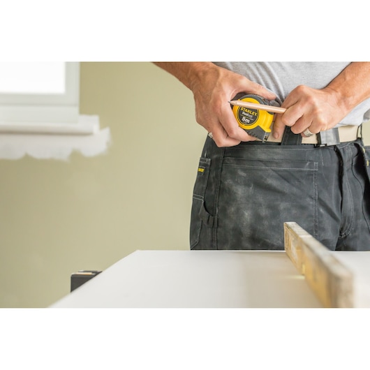 STANLEY STHT36803-0 rolmaat vastgehouden door werknemer in werkbroek naast witte tafel.