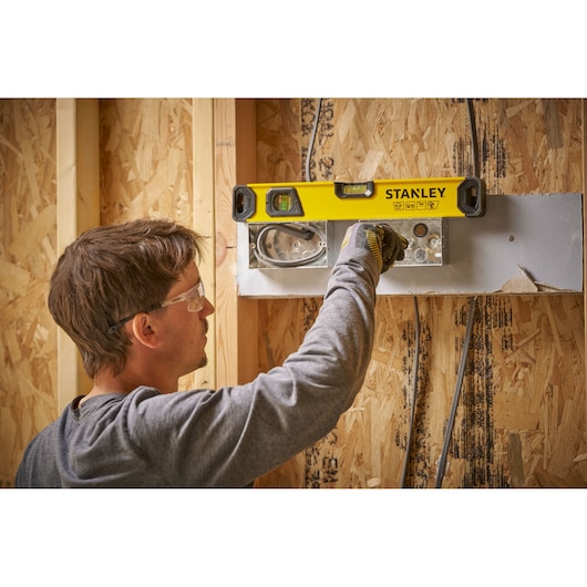 Een persoon gebruikt een STANLEY waterpas (model STHT41016-0) om de uitlijning van een metalen onderdeel op een houten muur in aanbouw te controleren.