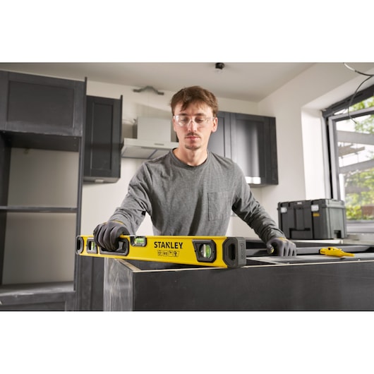 Een persoon met handschoenen gebruikt een gele STANLEY waterpas om de horizontale uitlijning van een keukenblad te controleren tijdens de installatie. De scène vindt plaats in een moderne keuken die in aanbouw is.