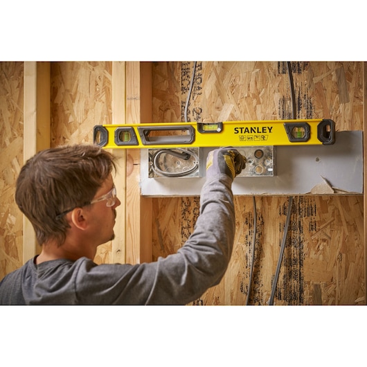 Een persoon gebruikt een gele STANLEY waterpas op een houten wandframe tijdens de bouw. Het gezicht van de persoon is vervaagd voor privacy. De waterpas wordt gebruikt om een elektrisch montageplaat waterpas te zetten.