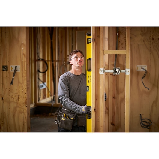 Een persoon met een vervaagd gezicht houdt een gele STANLEY waterpas vast in een houten frame van een huis in aanbouw. De persoon draagt een gereedschapsriem en handschoenen, en er zijn elektrische bedradingen zichtbaar in de muur.