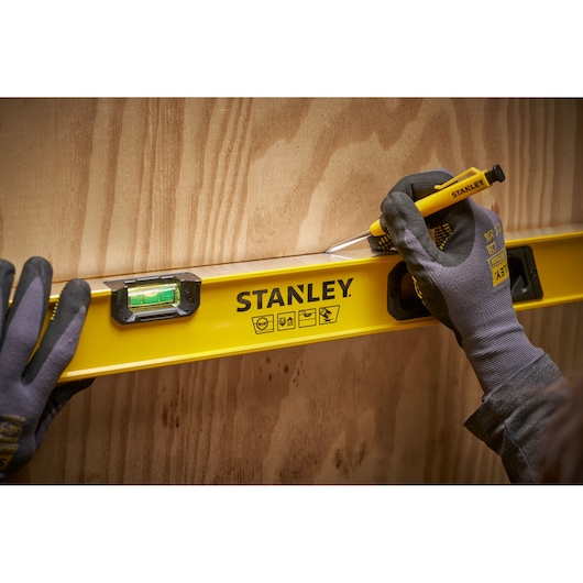 Een persoon met handschoenen gebruikt een gele STANLEY waterpas en een potlood om een lijn op een houten oppervlak te markeren.