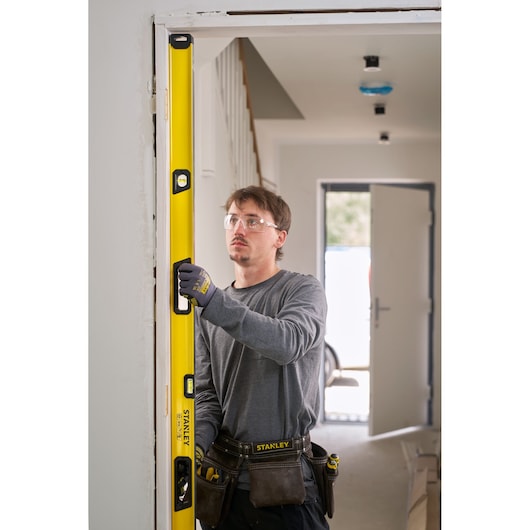 Een persoon met werkhandschoenen en een gereedschapsriem gebruikt een gele STANLEY waterpas om de verticale uitlijning van een deurkozijn in een huis in aanbouw of renovatie te controleren.