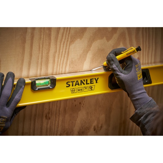 Een persoon met handschoenen gebruikt een gele STANLEY waterpas en een geel gereedschap om een rechte lijn op een houten oppervlak te markeren.