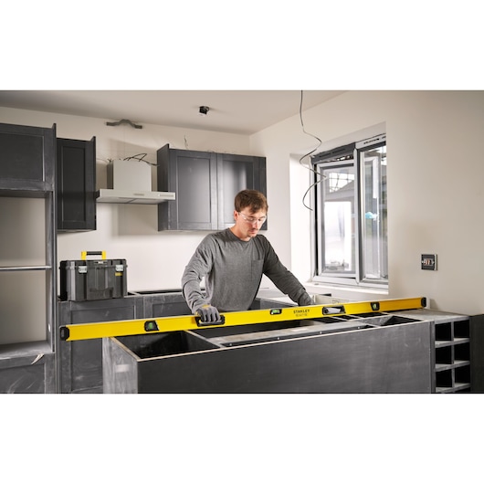 Een persoon gebruikt een gele STANLEY waterpas om oppervlakken uit te lijnen tijdens de installatie van een keuken. De persoon draagt handschoenen en bevindt zich in een moderne keuken in aanbouw.