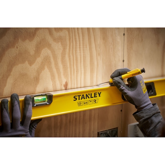 Een persoon met handschoenen gebruikt een gele STANLEY waterpas op een houten wand. De persoon houdt ook een gele schroevendraaier vast en markeert of positioneert langs de rand van de waterpas.