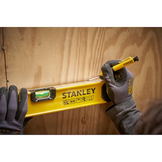 Een persoon met handschoenen gebruikt een Stanley waterpas en een markeergereedschap op een houten oppervlak. De Stanley waterpas is felgeel met zwarte markeringen en een groene libel.