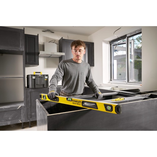 Persoon gebruikt een gele STANLEY waterpas (model STHT41736-0) in een keuken die in aanbouw is. De persoon draagt handschoenen en grijze kleding en stelt een aanrechtblad af. Bouwgereedschap en materialen zijn zichtbaar op de achtergrond.