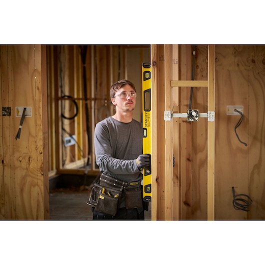 Een persoon met een vervaagd gezicht houdt een gele STANLEY waterpas tegen een houten wandframe op een bouwplaats. De persoon draagt werkhandschoenen en een gereedschapsriem met diverse gereedschappen.