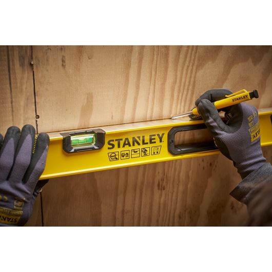 Een persoon met handschoenen gebruikt een gele STANLEY waterpas tegen een houten oppervlak en maakt een lijn met een STANLEY stanleymes.