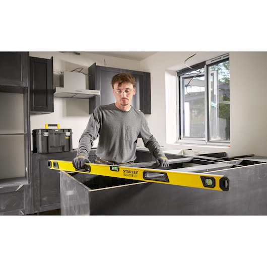 Persoon gebruikt een Stanley waterpas om een aanrechtblad uit te lijnen en te meten in een keuken in aanbouw, met gereedschapskisten en kasten zichtbaar op de achtergrond.