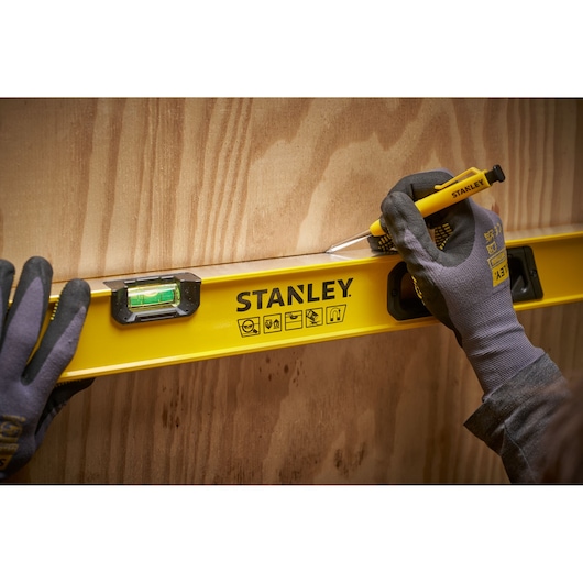 Een persoon met handschoenen gebruikt een gele STANLEY waterpas en een potlood om een rechte lijn op een houten oppervlak te markeren.