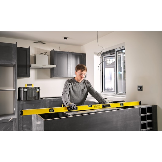Een persoon gebruikt een STANLEY waterpas (model STHT41772-0) om de uitlijning van een keukenblad te controleren tijdens de installatie. De persoon draagt handschoenen en werkt in een moderne keuken in aanbouw.