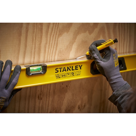 Een persoon met werkhandschoenen gebruikt een gele STANLEY waterpas tegen een houten oppervlak en tekent een lijn met een STANLEY gereedschap.