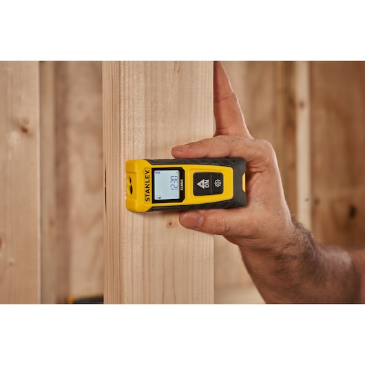 Télémètre laser STANLEY STHT77065-0 tenu contre une poutre en bois, écran allumé, en environnement de chantier.