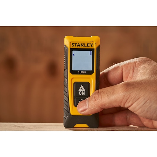 Photo du télémètre laser STANLEY SLM65 SKU STHT77065-0 en main, jaune et noir, écran visible.