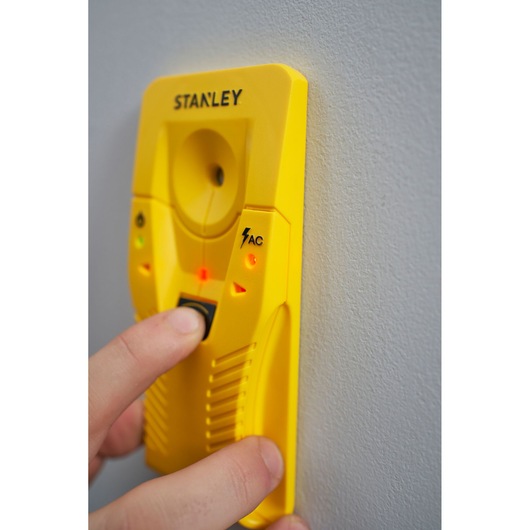STANLEY STHT77587-0 gele leidingzoeker met AC-detectie tegen een muur, hand drukt op knop.