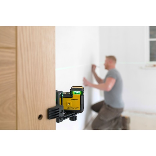 STANLEY STHT77594-1 kruislijnlaser bevestigd op hout, projecteert groene laserlijn, man werkt op de achtergrond.