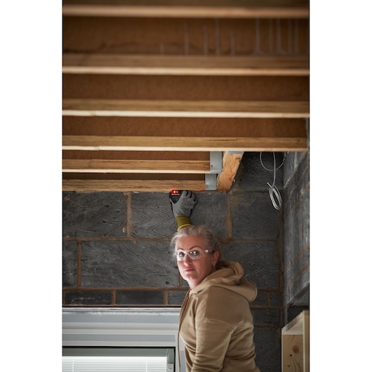 STANLEY STHT77666-0 outil de mesure laser utilisé par une personne gantée, pointant des poutres en bois à l'intérieur.
