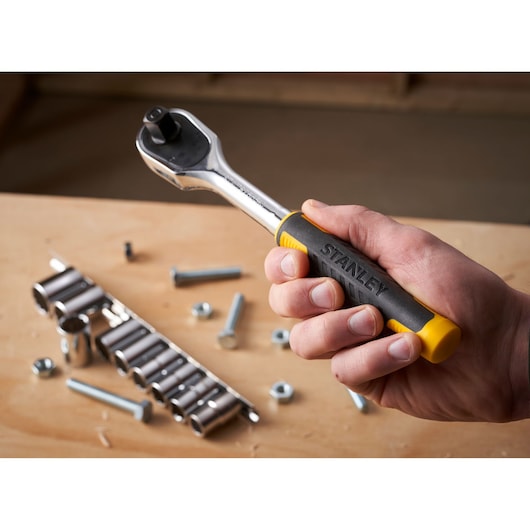 STANLEY STMT82671-0 ratelsleutel met zwart-gele handgreep, vastgehouden in hand, dopsleutelset op de achtergrond.