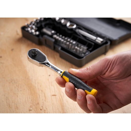 STANLEY STMT82672-0 ratelsleutel met zwart-gele grip in hand, gereedschapskist op de achtergrond.