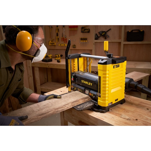 Photo du rabot STANLEY STP18-B2 utilisé pour aplanir une planche en bois dans un atelier, équipements de sécurité visibles.