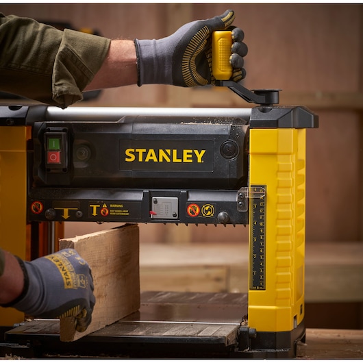 STANLEY STP18-B2 raboteuse à bois en utilisation, angle latéral, jaune et noir, mains gantées ajustant les commandes.