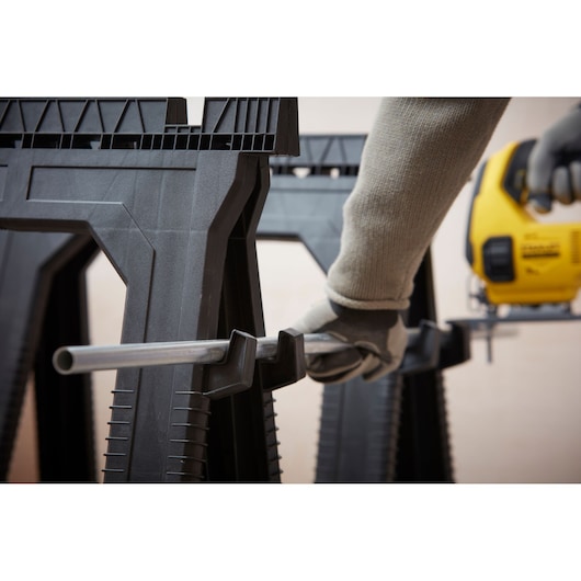 STANLEY STST1-70713 zaagbok close-up met gehandschoende hand die metalen buis vasthoudt, DEWALT decoupeerzaag op achtergrond.