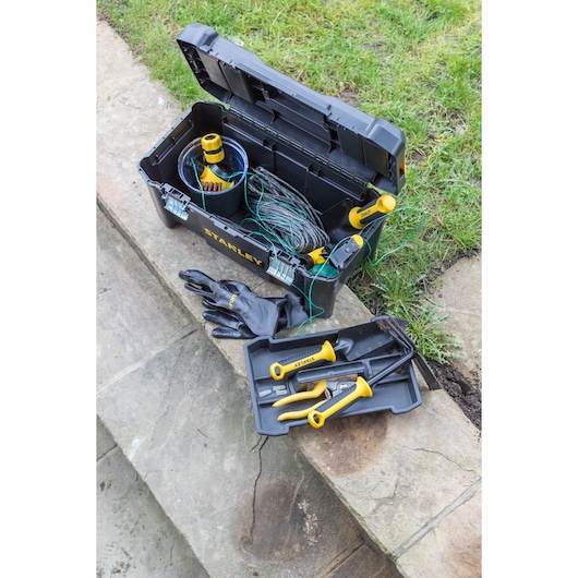Foto van STANLEY STST1-75521 zwarte gereedschapskist met geelhandvat tuingereedschap en handschoenen buiten.