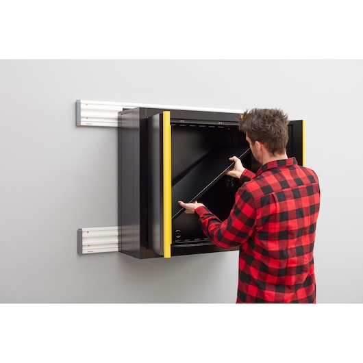 STANLEY STST97598-1 wandgemonteerde zwarte opbergkast met gele rand, persoon plaatst plank, zijaanzicht.