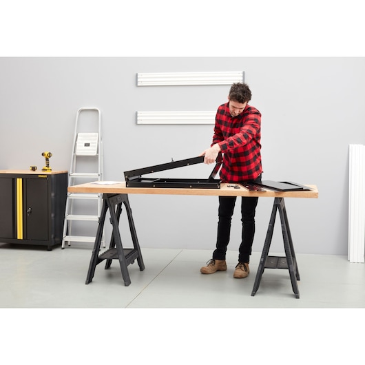 Photo de l'établi STANLEY STST97599-1 dans un atelier, personne assemblant un cadre métallique noir sur une surface en bois.