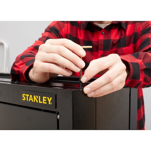 Photo de mains assemblant le coffre à outils STANLEY STST97599-1 avec clé hexagonale, gros plan sur le logo et la vis.